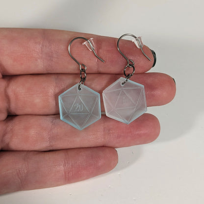 A pair of frosty blue and white dangly, flat d20 earrings with silver color titanium and niobium hardware with bell shaped rubber backs held on on a hand on a white background. The earring on the left has a 20 on it and the earring on the right has a 1 on it, both in a fantasy style font.