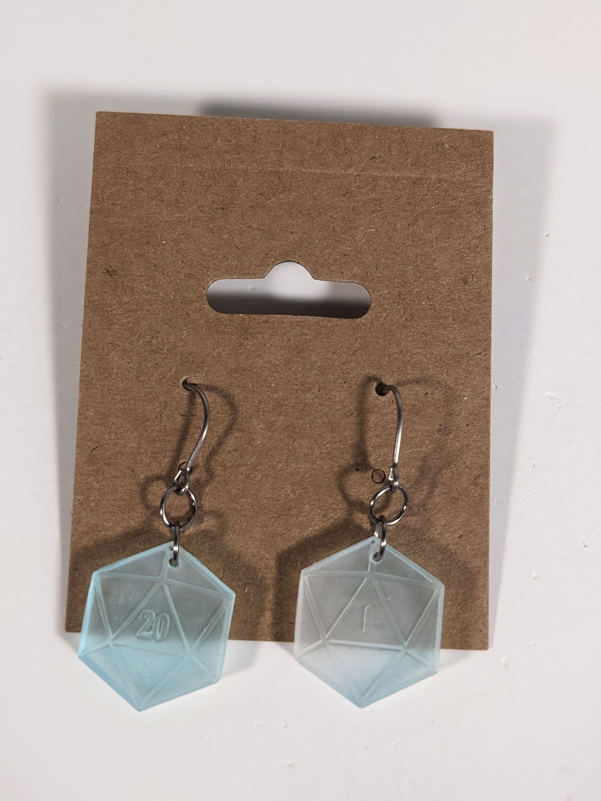 A pair of frosty blue and white dangly, flat d20 earrings with silver color titanium and niobium hardware on a kraft earring card on a white background. The earring on the left has a 20 on it and the earring on the right has a 1 on it, both in a fantasy style font.