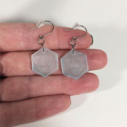A pair of frosty blue and white dangly, flat d20 earrings with silver color titanium and niobium hardware with bell shaped rubber backs held on on a hand on a white background.