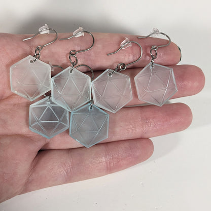 Three pairs of frosty blue and white dangly, flat d20 earrings with silver color titanium and niobium hardware with bell shaped rubber backs held on on a hand on a white background.