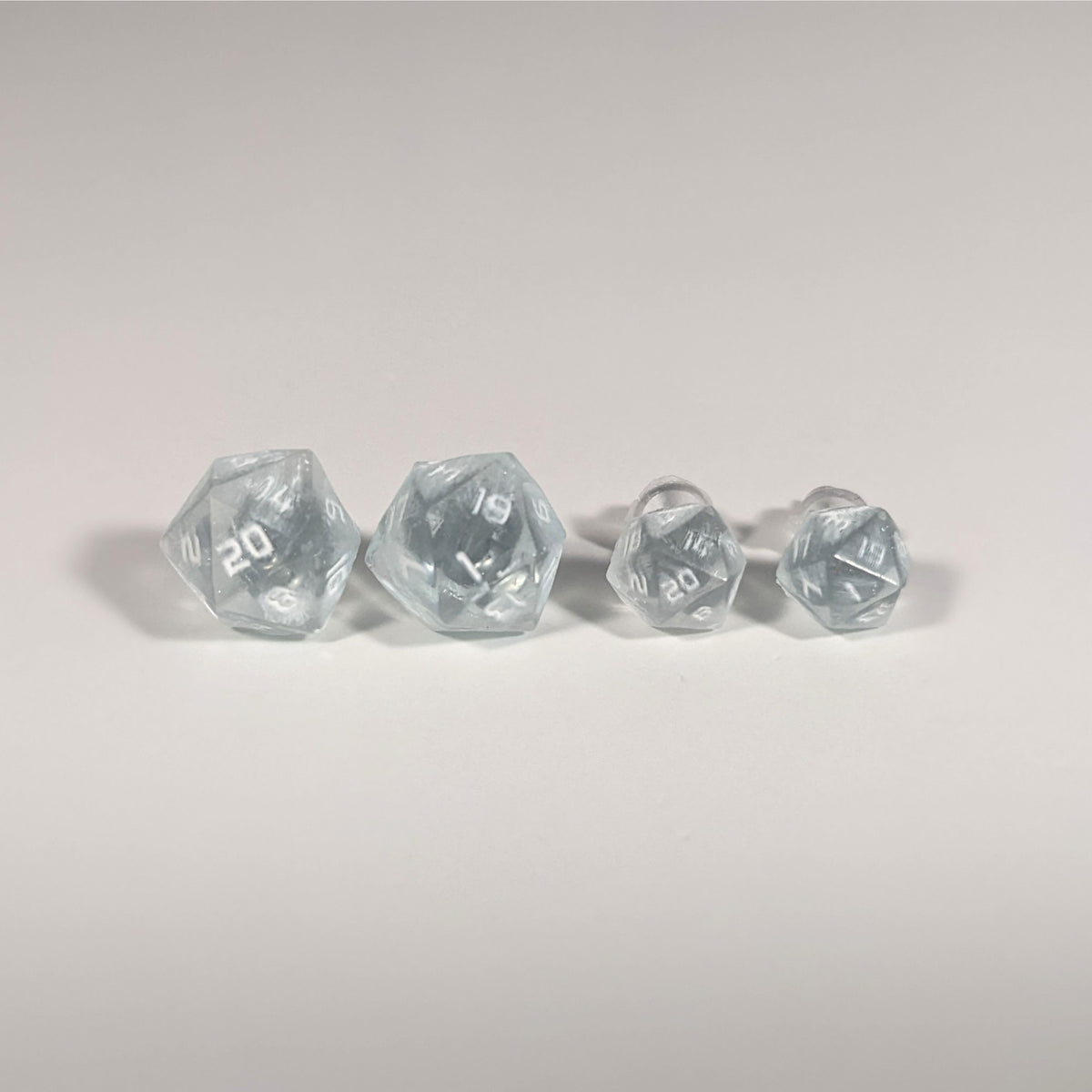 Two pairs of handmade d20 earrings with titanium hardware that are frosty blue and white with white numbers on a white background sit in a line with the larger pair on the left and the smaller pair on the right. The earrings on the left of each pair centers the 20 face and the earrings on the right of each pair centers the 1 face.