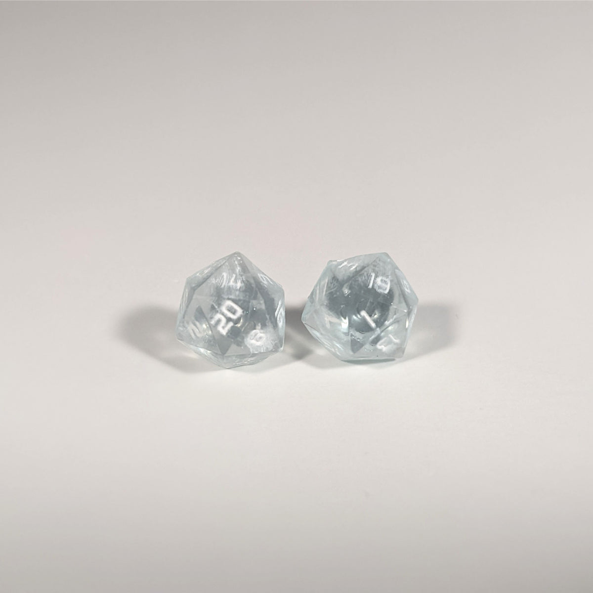 A single pair of the larger size handmade d20 earrings with titanium hardware that are frosty blue and white with white numbers on a white background. The earring on the left centers the 20 face and the earring on the right centers the 1 face.
