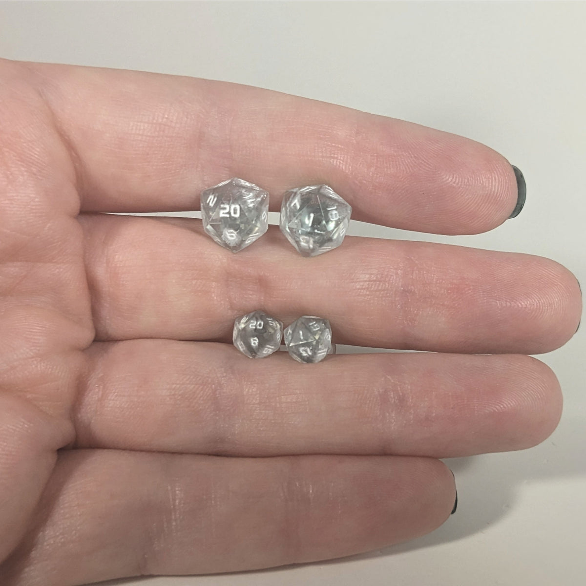 Two pairs of handmade d20 earrings with titanium hardware that are frosty blue and white with white numbers held between the fingers of a flat hand. The larger pair is between the pointer and middle finger, the smaller between the middle and ring finger. The earrings on the left of each pair centers the 20 face and the earrings on the right of each pair centers the 1 face.