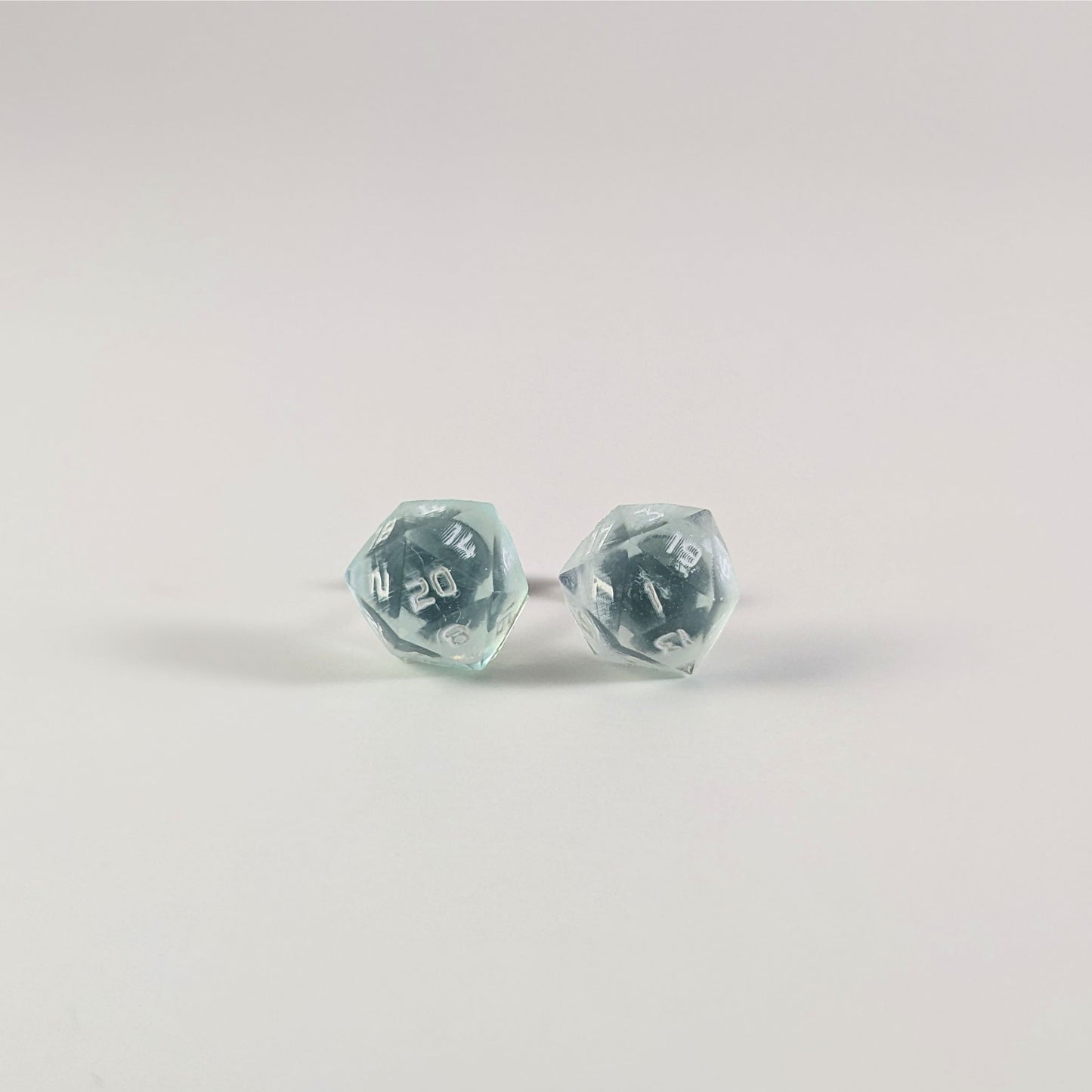 A single pair of the larger size handmade d20 earrings that are transparent white with hints of glittery blue and opaque white numbers on a white background. The earring on the left centers the 20 face and the earring on the right centers the 1 face.