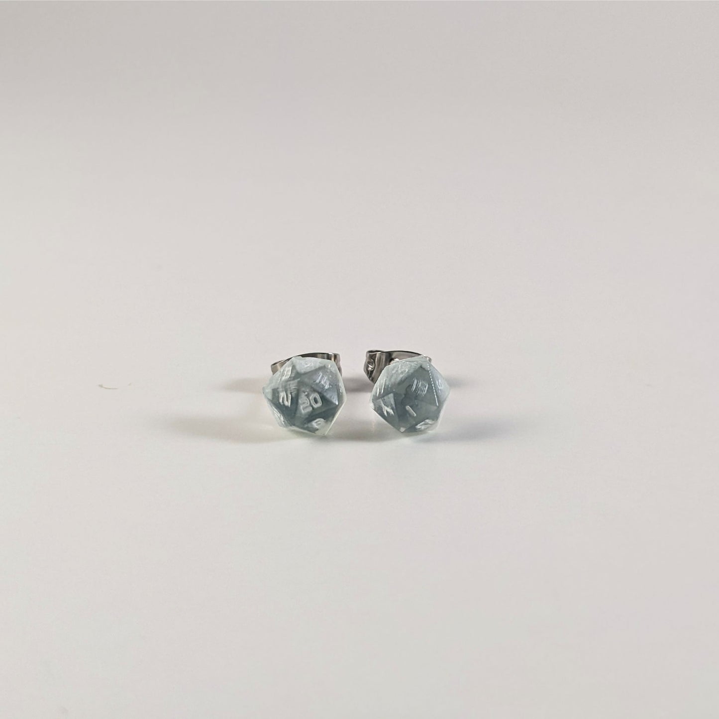A single pair of the smaller size handmade d20 earrings that are transparent white with hints of glittery blue and opaque white numbers on a white background. The earring on the left centers the 20 face and the earring on the right centers the 1 face.