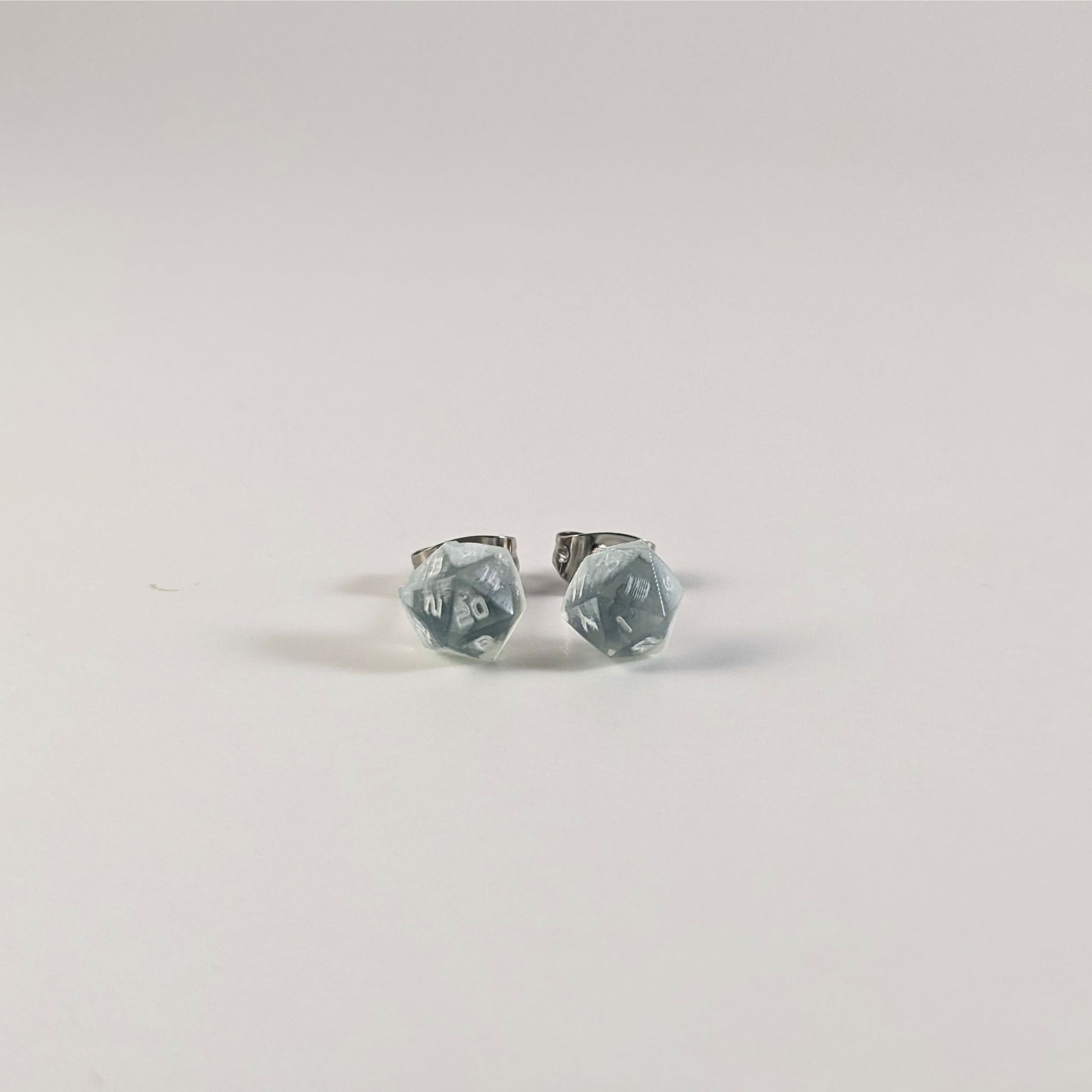 A single pair of the smaller size handmade d20 earrings that are transparent white with hints of glittery blue and opaque white numbers on a white background. The earring on the left centers the 20 face and the earring on the right centers the 1 face.