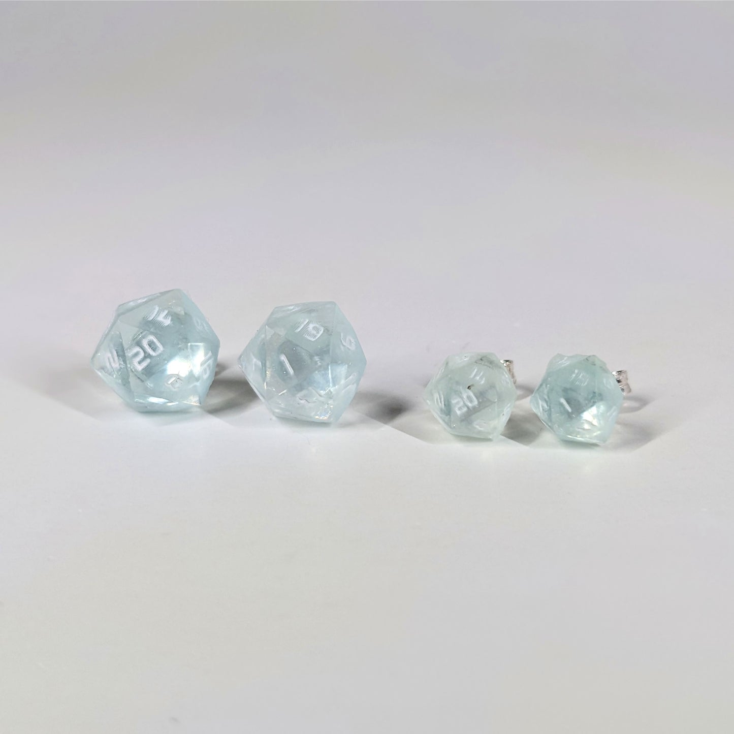 Two pairs of handmade d20 earrings with sterling silver hardware that are transparent white with hints of glittery blue and opaque white numbers on a white background sit in a line with the larger pair on the left and the smaller pair on the right. The earrings on the left of each pair centers the 20 face and the earrings on the right of each pair centers the 1 face.