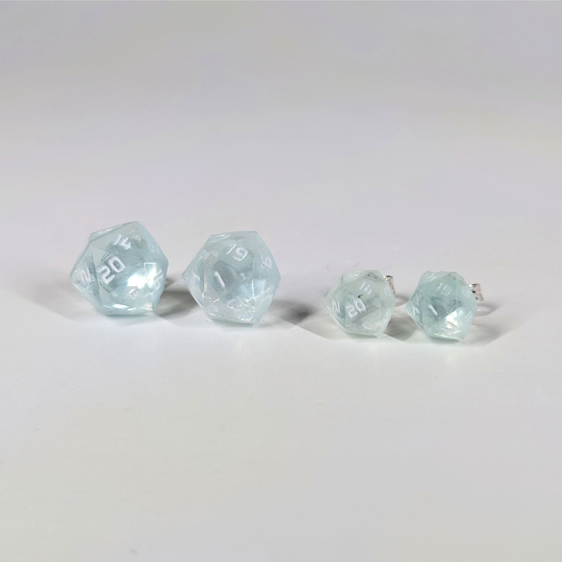 Two pairs of handmade d20 earrings with sterling silver hardware that are transparent white with hints of glittery blue and opaque white numbers on a white background sit in a line with the larger pair on the left and the smaller pair on the right. The earrings on the left of each pair centers the 20 face and the earrings on the right of each pair centers the 1 face.