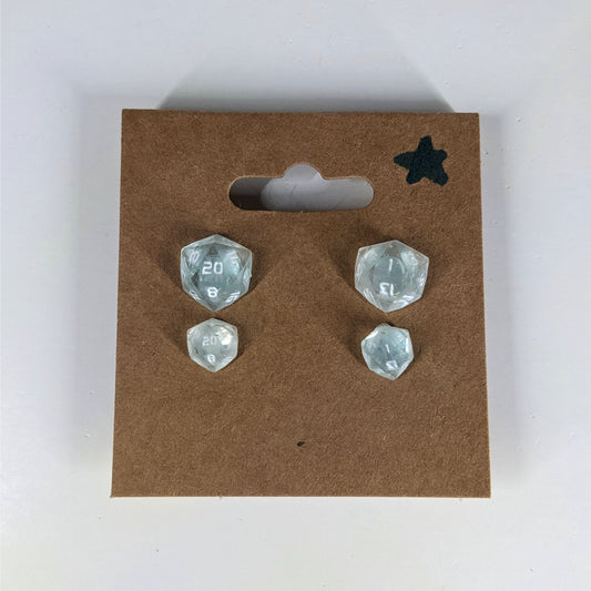 Two pairs of handmade d20 earrings with sterling silver hardware that are transparent white with hints if glittery blue with opaque numbers on a kraft brown earring card with a star in the top right corner. The earrings on the left of each pair centers the 20 face and the earrings on the right of each pair centers the 1 face.