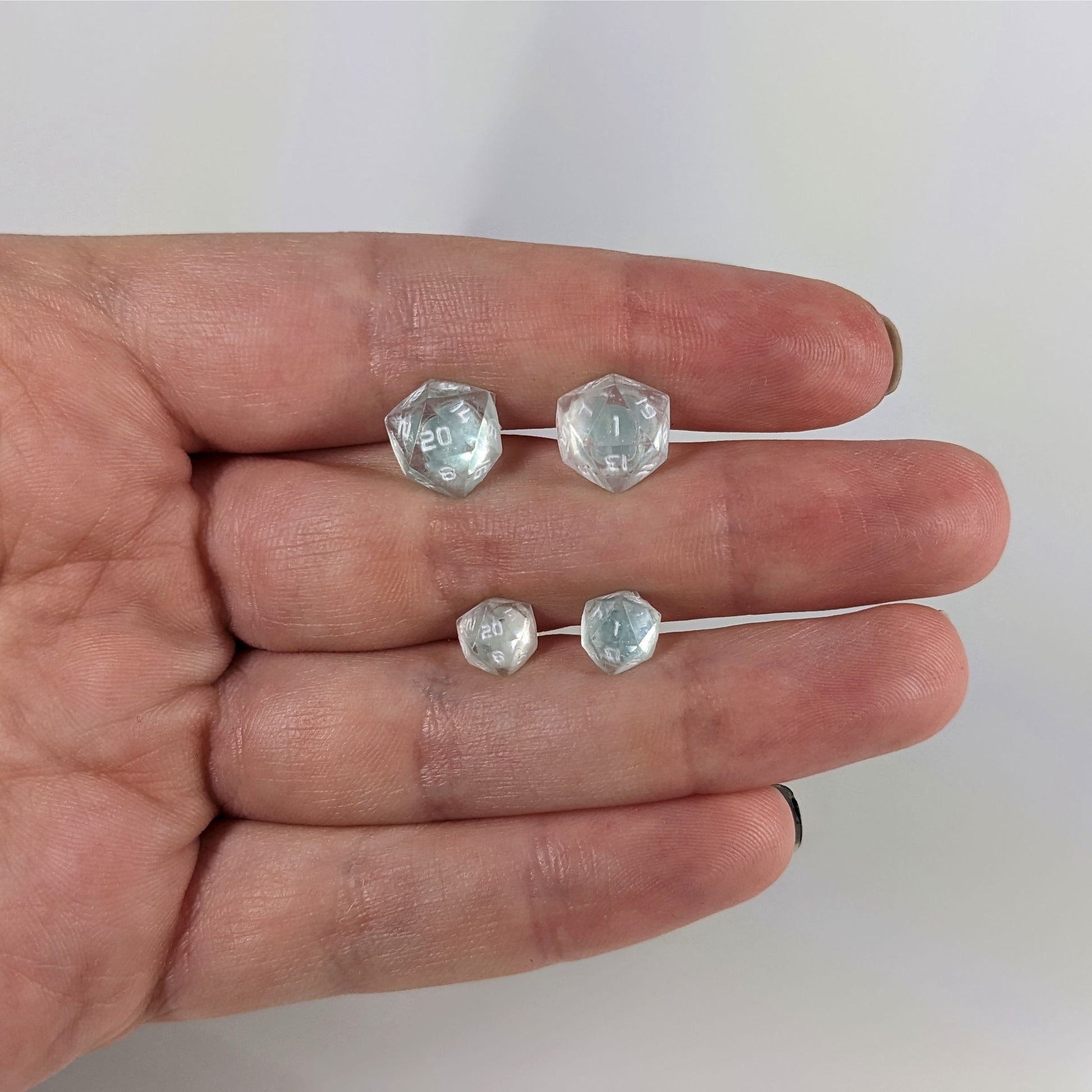 Two pairs of handmade d20 earrings with sterling silver hardware that are transparent white with hints of glittery blue and opaque white numbers held between the fingers of a flat hand. The larger pair is between the pointer and middle finger, the smaller between the middle and ring finger. The earrings on the left of each pair centers the 20 face and the earrings on the right of each pair centers the 1 face.