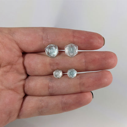 Two pairs of handmade d20 earrings with sterling silver hardware that are transparent white with hints of glittery blue and opaque white numbers held between the fingers of a flat hand. The larger pair is between the pointer and middle finger, the smaller between the middle and ring finger. The earrings on the left of each pair centers the 20 face and the earrings on the right of each pair centers the 1 face.