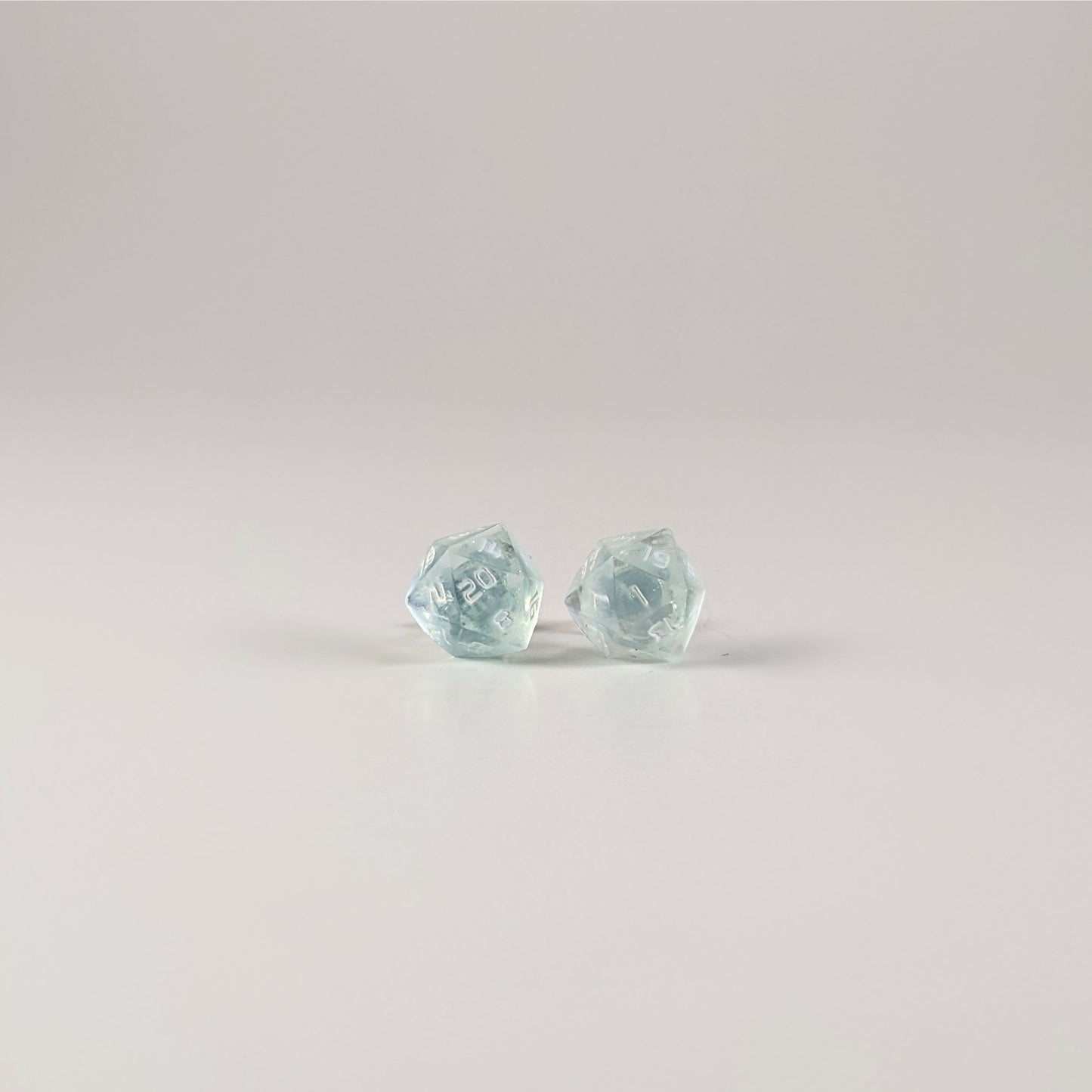 A single pair of the larger size handmade d20 earrings with sterling silver hardware that are transparent white with hints of glittery blue with opaque white numbers on a white background. The earring on the left centers the 20 face and the earring on the right centers the 1 face.