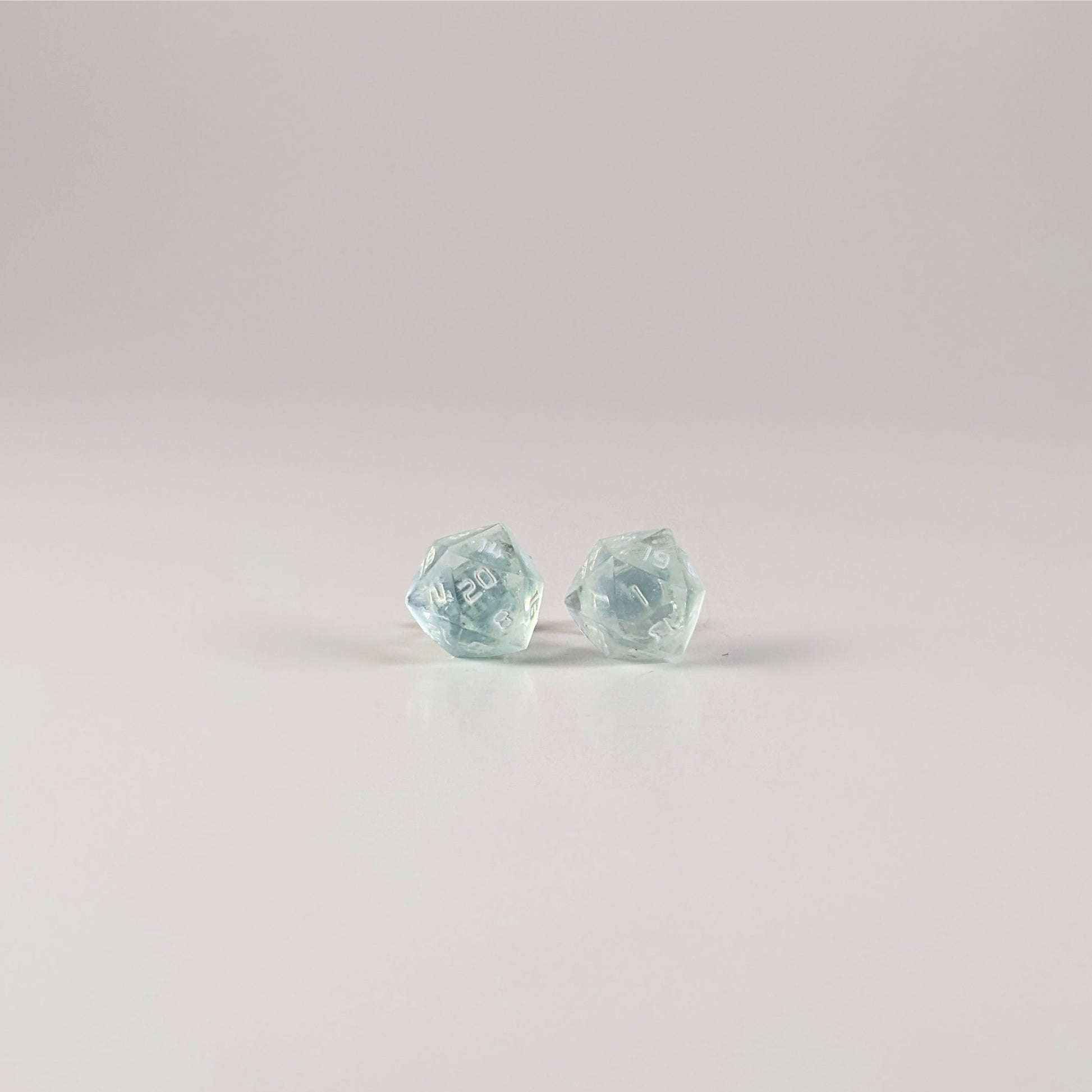 A single pair of the larger size handmade d20 earrings with sterling silver hardware that are transparent white with hints of glittery blue with opaque white numbers on a white background. The earring on the left centers the 20 face and the earring on the right centers the 1 face.