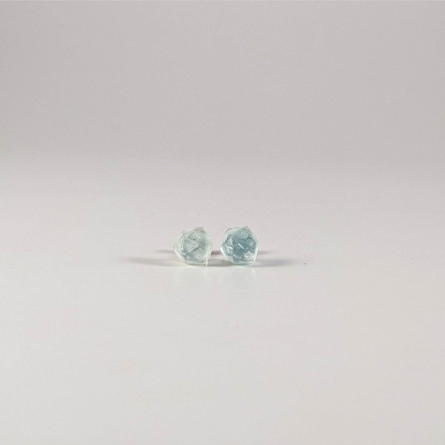 A single pair of the smaller size handmade d20 earrings with sterling silver hardware that are transparent white and hints of glittery blue with opaque white numbers on a white background. The earring on the left centers the 20 face and the earring on the right centers the 1 face.