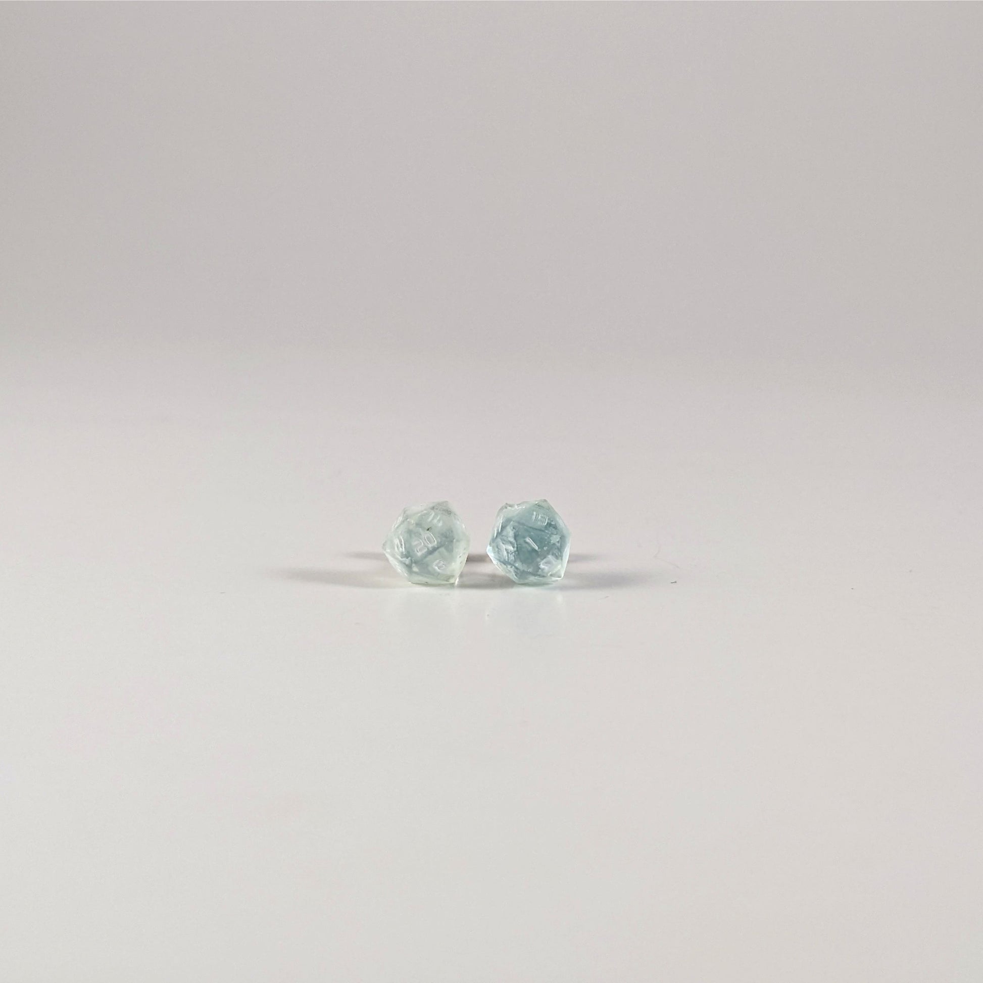 A single pair of the smaller size handmade d20 earrings with sterling silver hardware that are transparent white and hints of glittery blue with opaque white numbers on a white background. The earring on the left centers the 20 face and the earring on the right centers the 1 face.