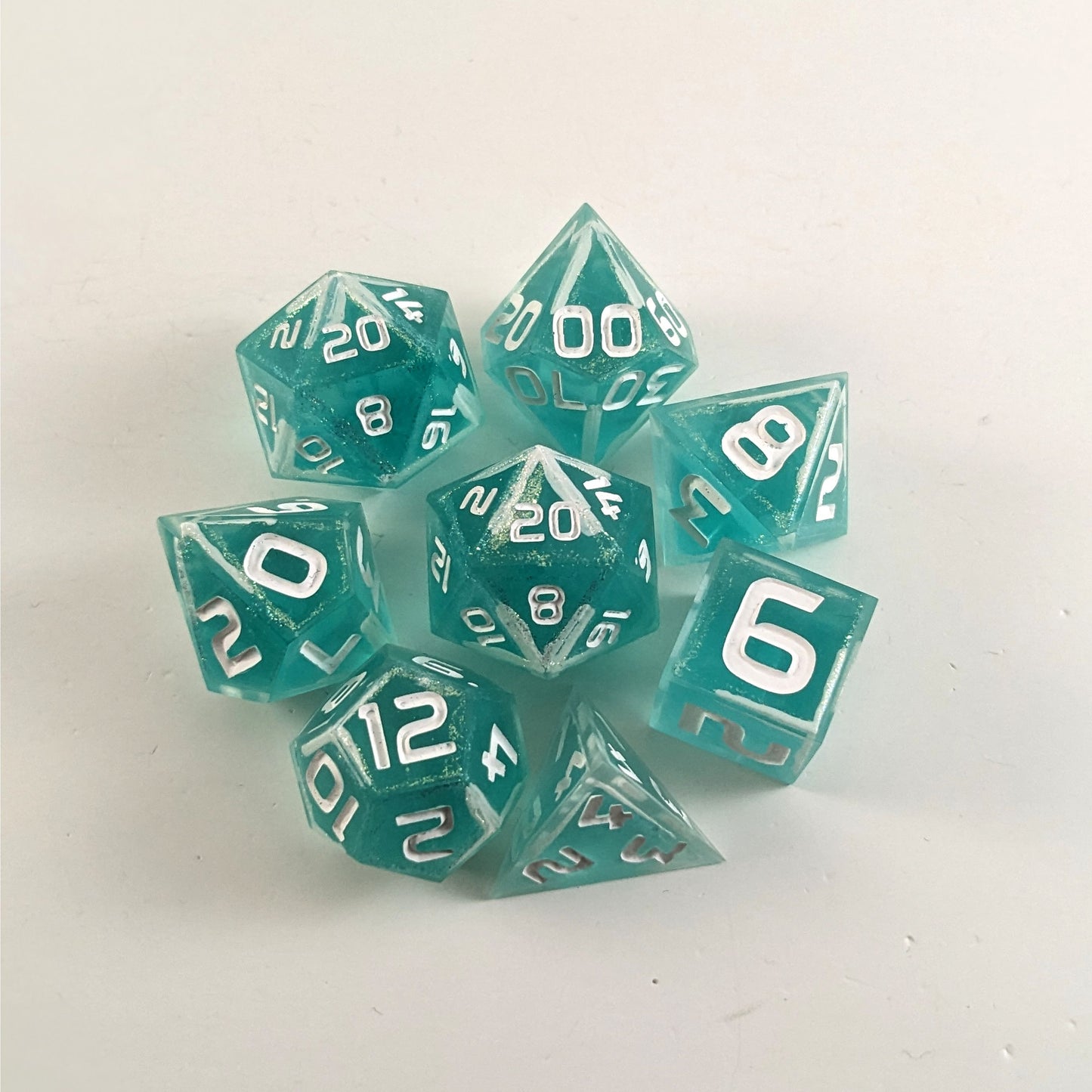 A complete set of 8 handmade artisan dice that are icy blue with edges painted with white and iridescent glitter in a clear shell with white numbers. The DnD dice set is arranged in a circle and features 2 D20s, one of which is in the center surrounded by the other 7 dice.