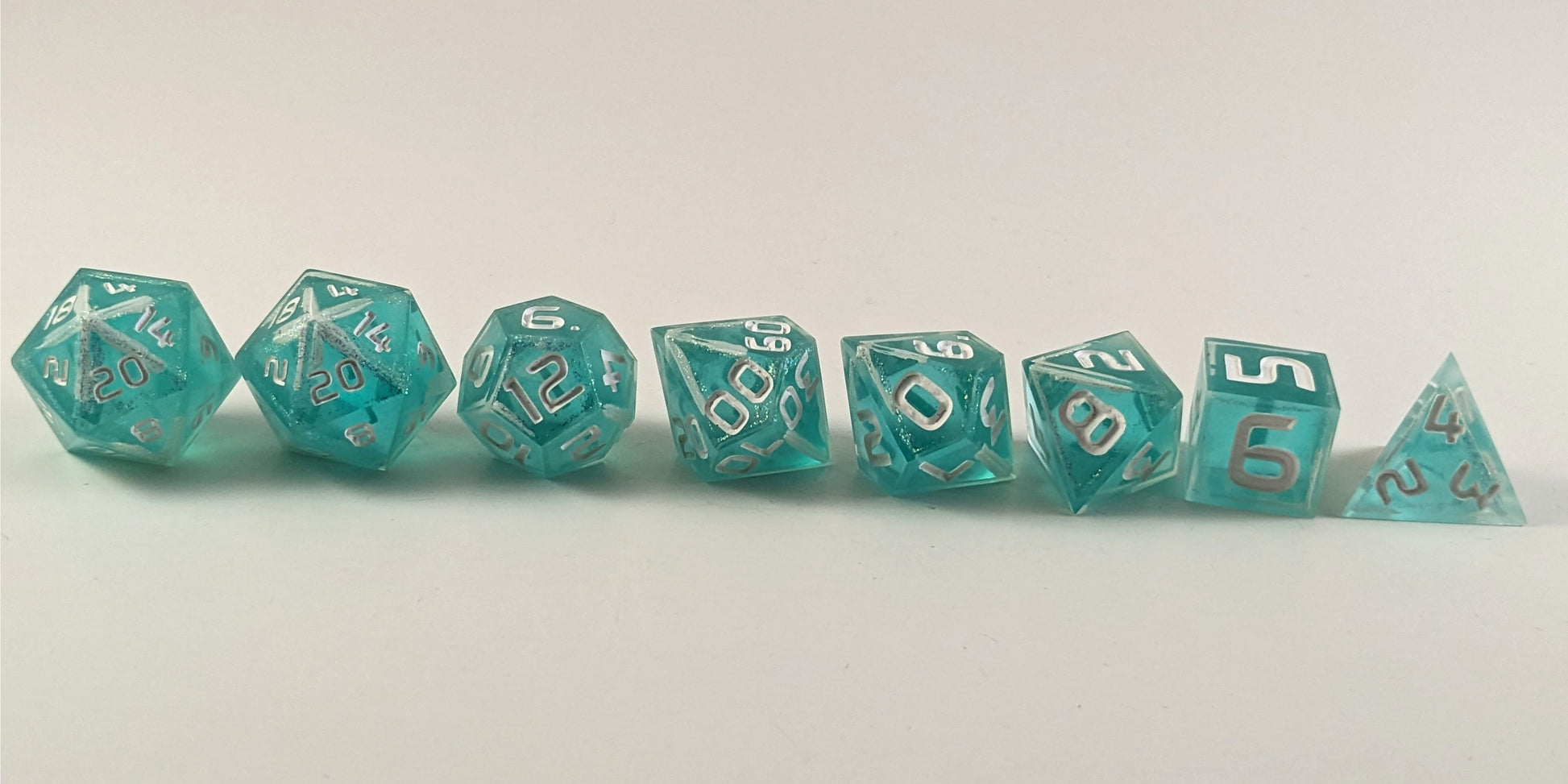 A straight on photo of a complete set of polyhedral dice that are icy blue with edges painted with white and iridescent glitter in a clear shell with white numbers arranged in a straight line. They are lined up with the 2 d20s on the left going down to the d4 on the right side. The highest value number of each die is facing forward.