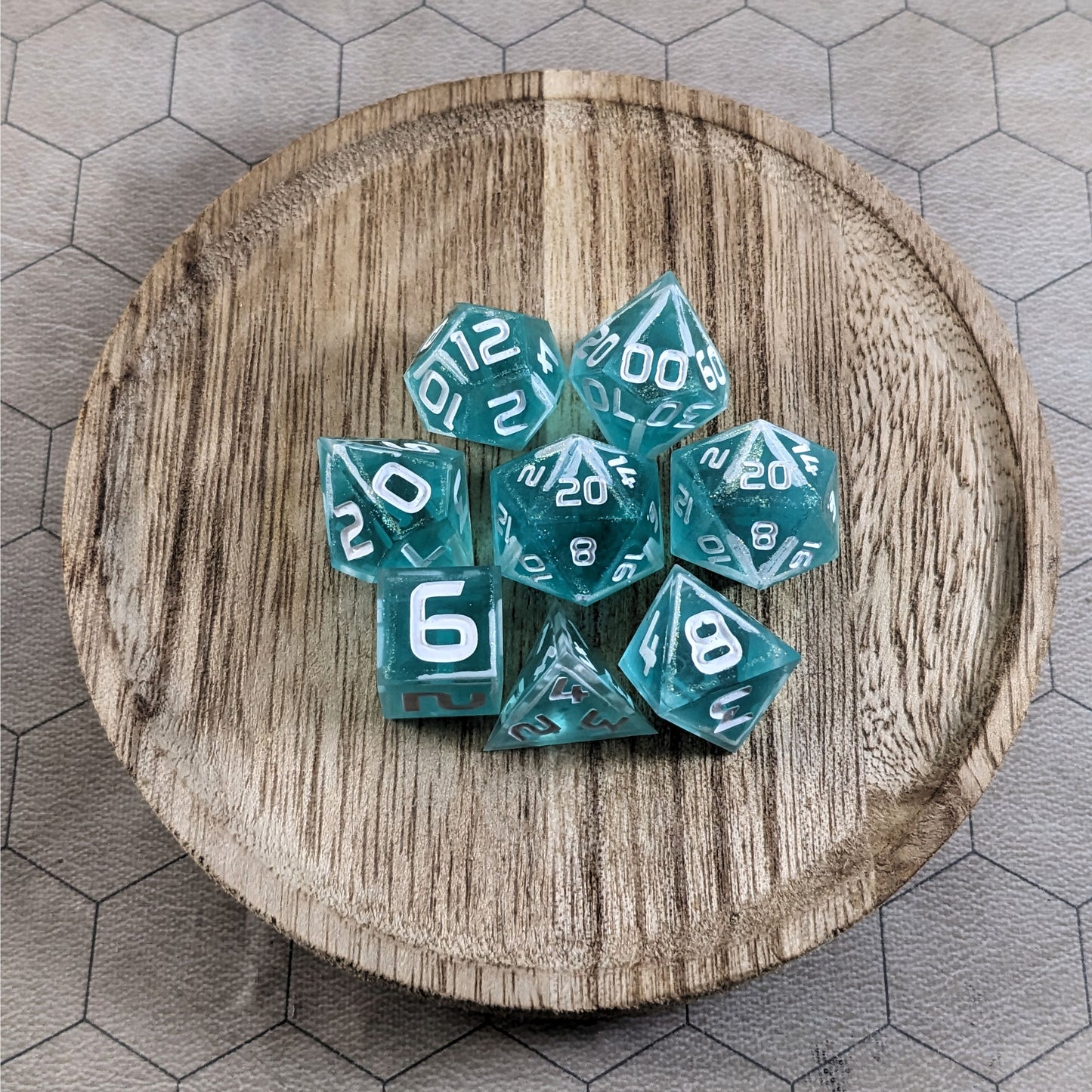 A complete set of 8 handmade artisan dice that are icy blue with edges painted with white and iridescent glitter in a clear shell with white numbers. The DnD dice set is on a wooden circle and the dice are arranged in a circle and features 2 D20s, one of which is in the center surrounded by the other 7 dice.