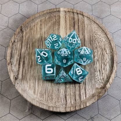 A complete set of 8 handmade artisan dice that are icy blue with edges painted with white and iridescent glitter in a clear shell with white numbers. The DnD dice set is on a wooden circle and the dice are arranged in a circle and features 2 D20s, one of which is in the center surrounded by the other 7 dice.
