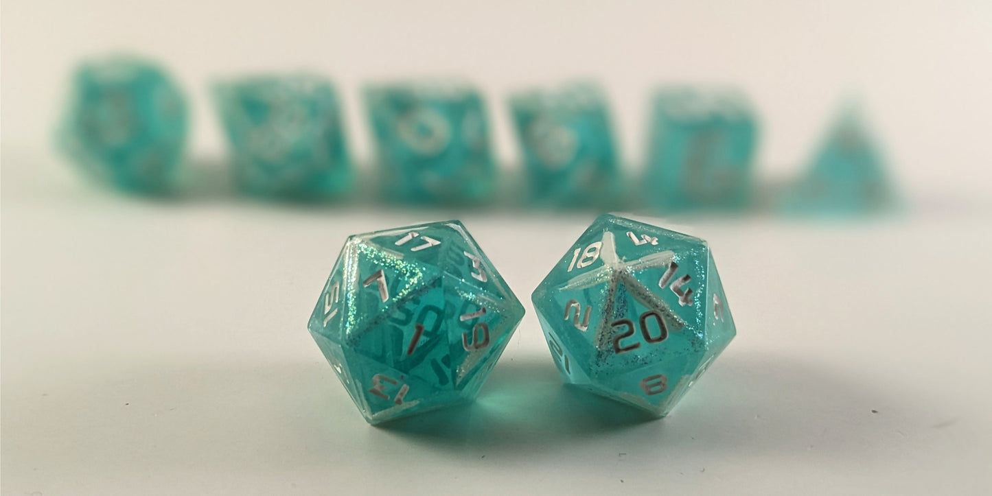 A straight on photo of a standard set of dice that are icy blue with edges painted with white and iridescent glitter in a clear shell with white numbers focusing on 2 d20s in the center. The d20 on the left has the 1 face centered and the d20 on the right has the 20 face centered. The other 6 dice are lined up in the background but are out of focus, while the d20s are in sharp focus.