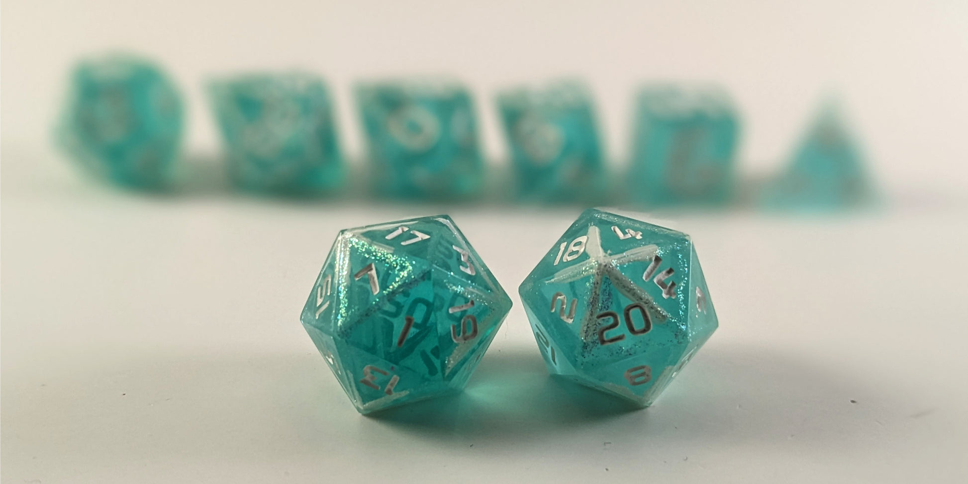 A straight on photo of a standard set of dice that are icy blue with edges painted with white and iridescent glitter in a clear shell with white numbers focusing on 2 d20s in the center. The d20 on the left has the 1 face centered and the d20 on the right has the 20 face centered. The other 6 dice are lined up in the background but are out of focus, while the d20s are in sharp focus.