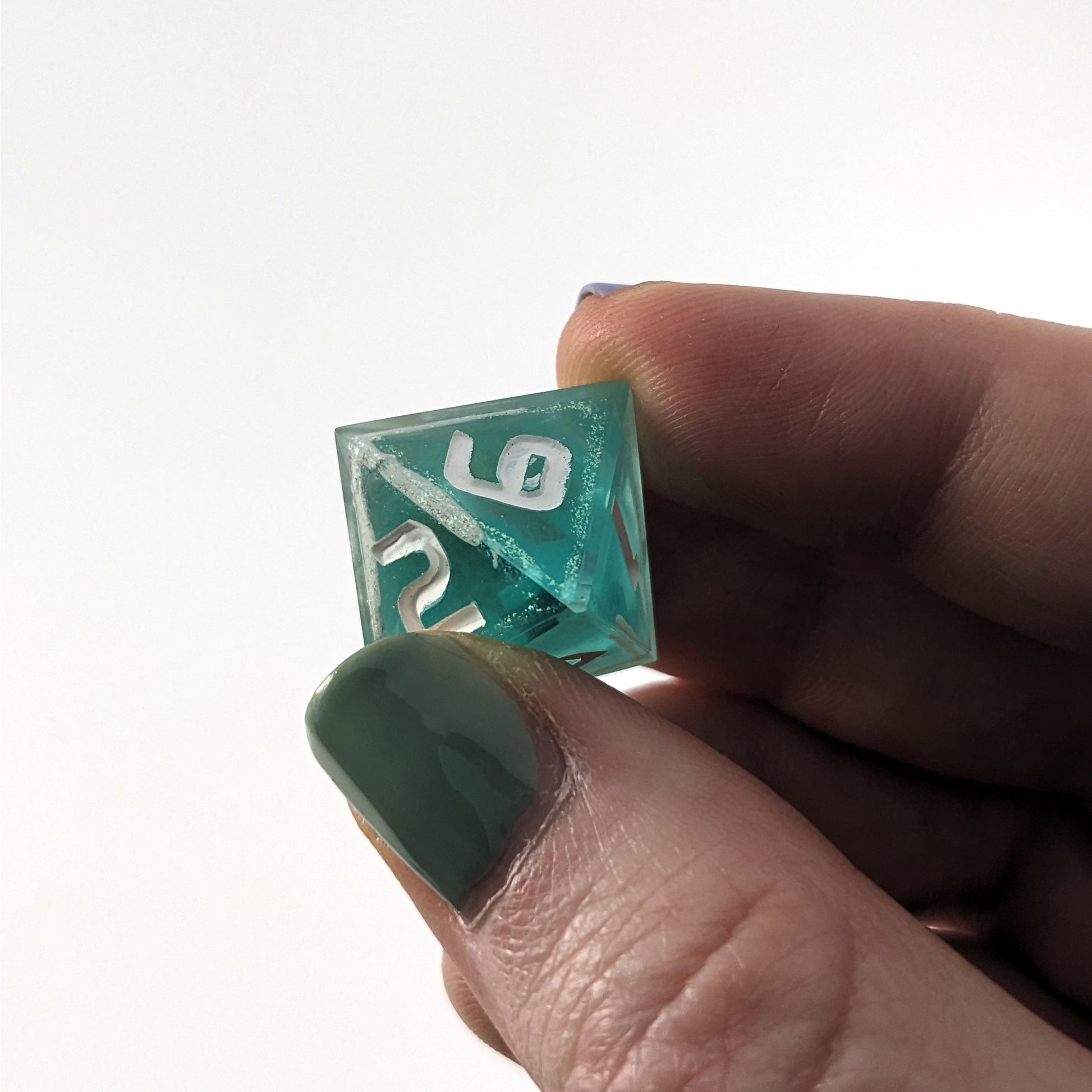 The d8 that is icy blue with edges painted with white and iridescent glitter in a clear shell with white is held up to the camera. The angle shows a small cosmetic flaw in the hole in the loop of the 6. The 6 is still clearly legible and the flaw would not affect rolling.