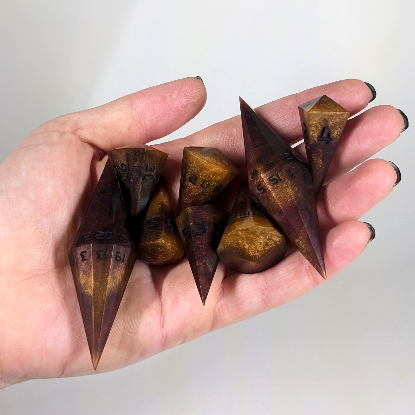 An 8 dice set of dagger style dice that are bright orange tempered with deep red like cooling lava with obsidian black numbers are held in a hand. The image shows that the set just fits in a hand, making them overall larger than a standard set of dnd dice.