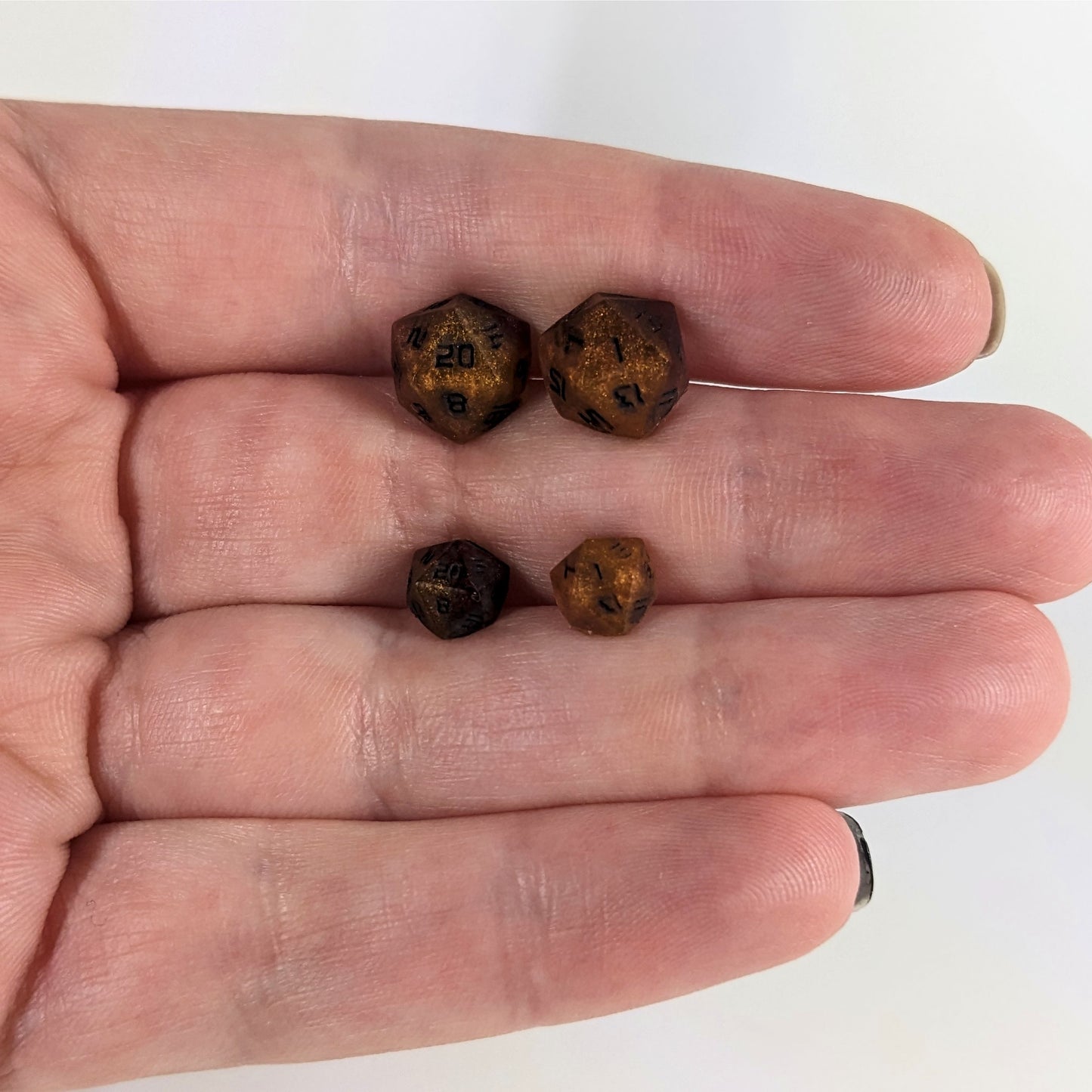 Two pairs of handmade d20 earrings that are orange and deep red with black numbers held between the fingers of a flat hand. The larger pair is between the pointer and middle finger, the smaller between the middle and ring finger. The earrings on the left of each pair centers the 20 face and the earrings on the right of each pair centers the 1 face.