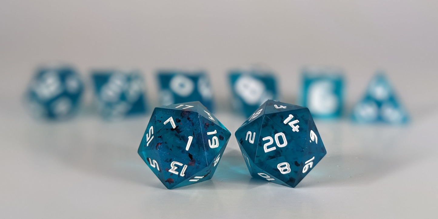 A straight on photo of a standard set of DnD dice that are blue with red flakies and white numbers focusing on 2 d20s in the center. The d20 on the left has the 1 face centered and the d20 on the right has the 20 face centered. The other 6 dice are lined up in the background but are out of focus, while the d20s are in sharp focus.
