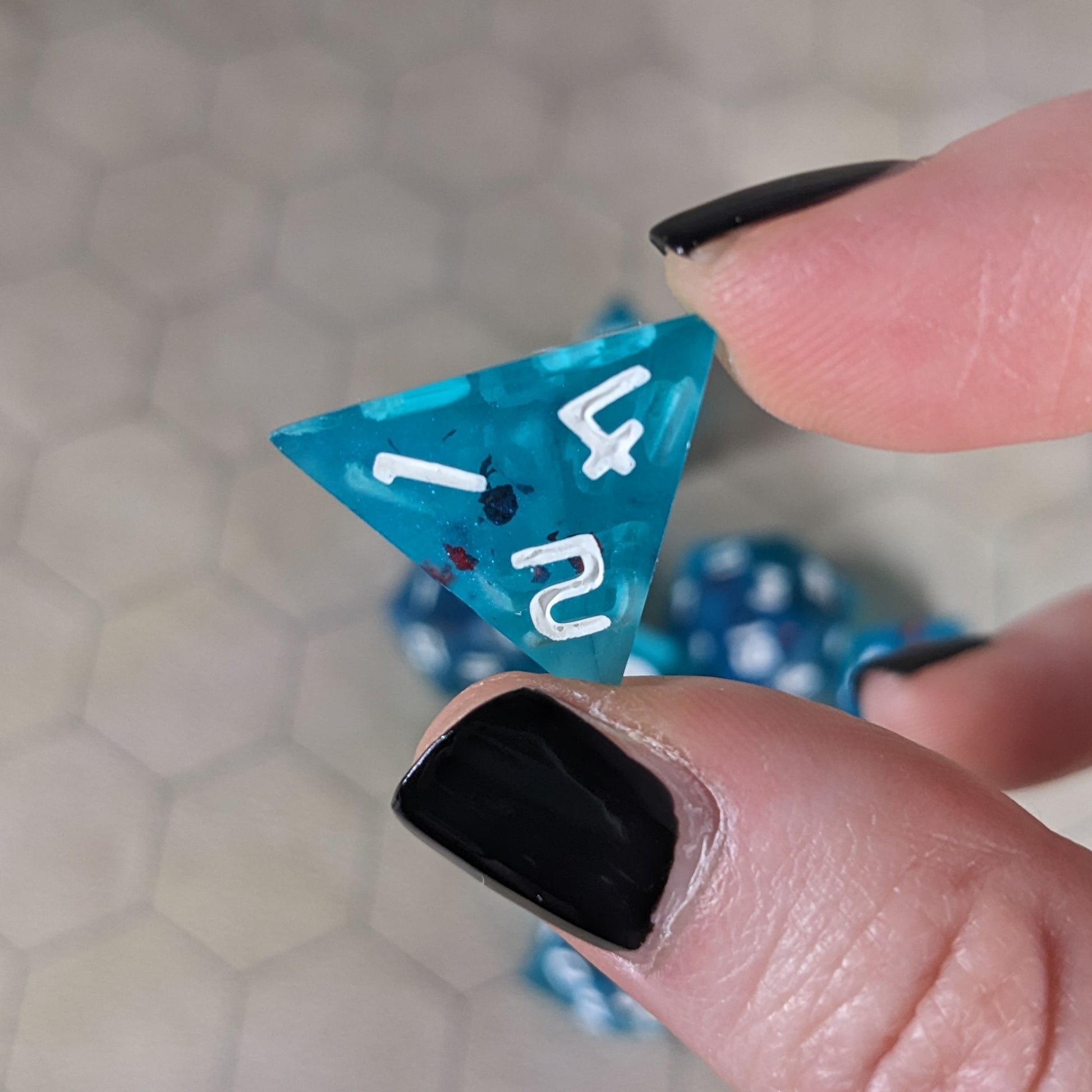 A d4 that is blue with red flakies and white numbers is held up to the camera to show off the red flakies suspended in the blue die. The 1, 2, 4 face is angled toward the camera.