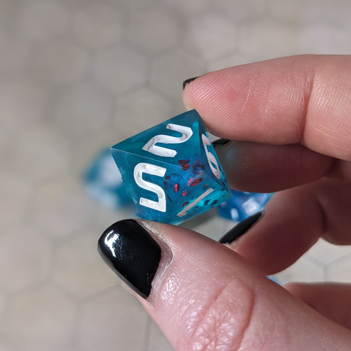 A d8 that is blue with red flakies and white numbers is held up to the camera to show off the red flakies suspended in the blue die. The 2 and 5 faces are angled toward the camera.