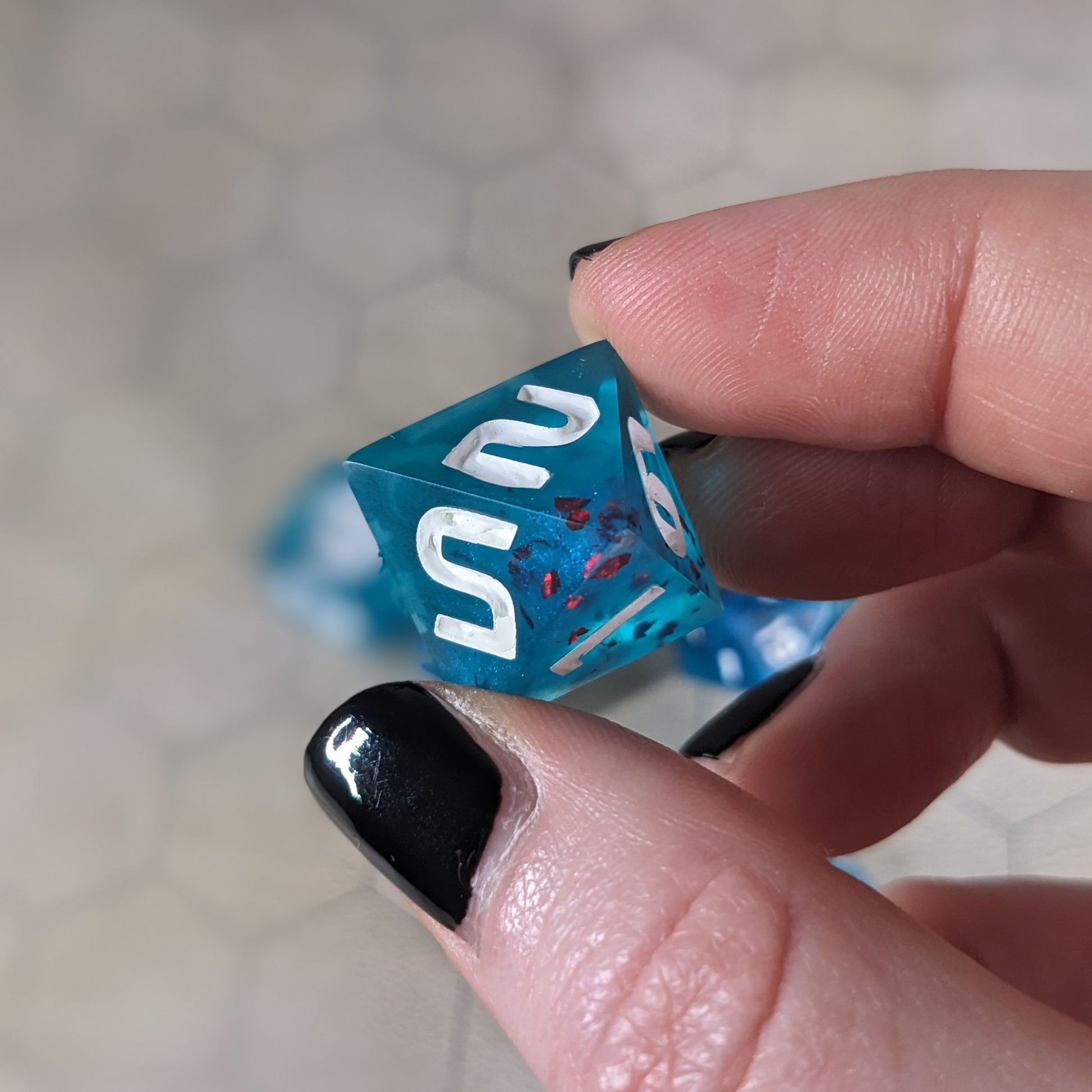 A d8 that is blue with red flakies and white numbers is held up to the camera to show off the red flakies suspended in the blue die. The 2 and 5 faces are angled toward the camera.