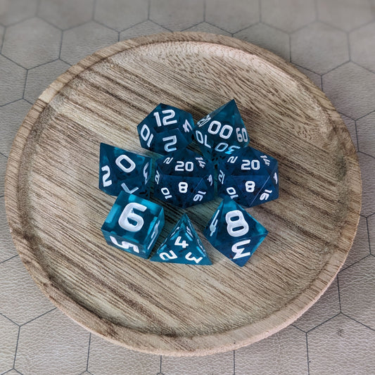  A complete set of 8 handmade artisan dice that are blue with red flakies and white numbers. The DnD dice set is on a wooded circle and the dice are arranged in a circle and features 2 D20s, one of which is in the center surrounded by the other 7 dice. 