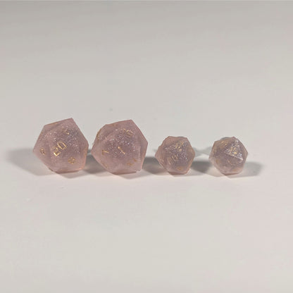 Two pairs of handmade d20 earrings with titanium hardware that are pastel pink with gold numbers on a white background sit in a line with the larger pair on the left and the smaller pair on the right. The earrings on the left of each pair centers the 20 face and the earrings on the right of each pair centers the 1 face.