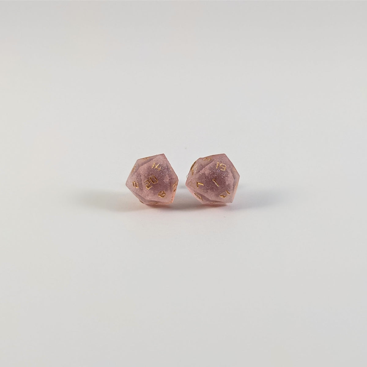 A single pair of the larger size handmade d20 earrings with titanium hardware that are pastel pink with gold numbers on a white background. The earring on the left centers the 20 face and the earring on the right centers the 1 face.