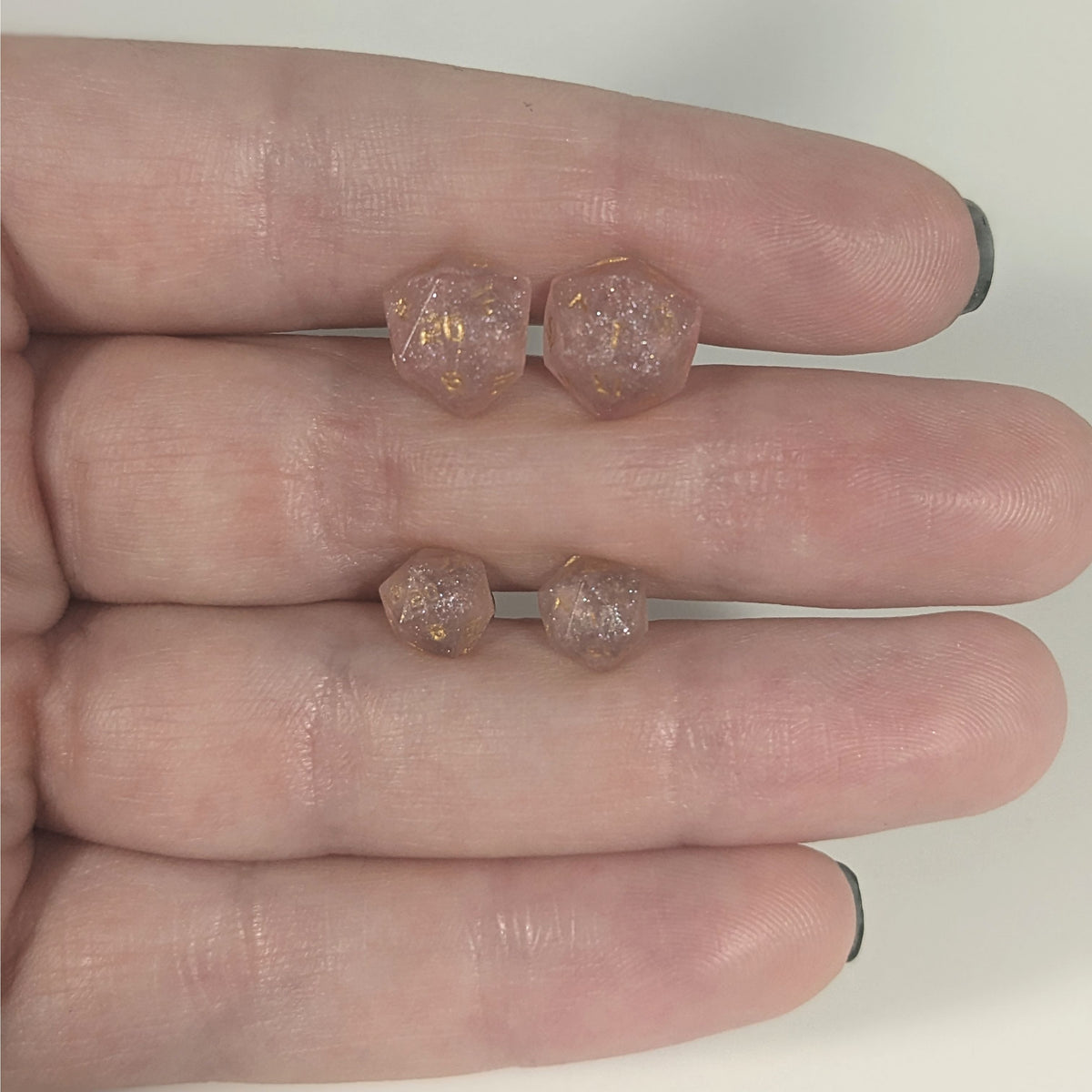 Two pairs of handmade d20 earrings with titanium hardware that are pastel pink with gold numbers held between the fingers of a flat hand. The larger pair is between the pointer and middle finger, the smaller between the middle and ring finger. The earrings on the left of each pair centers the 20 face and the earrings on the right of each pair centers the 1 face.