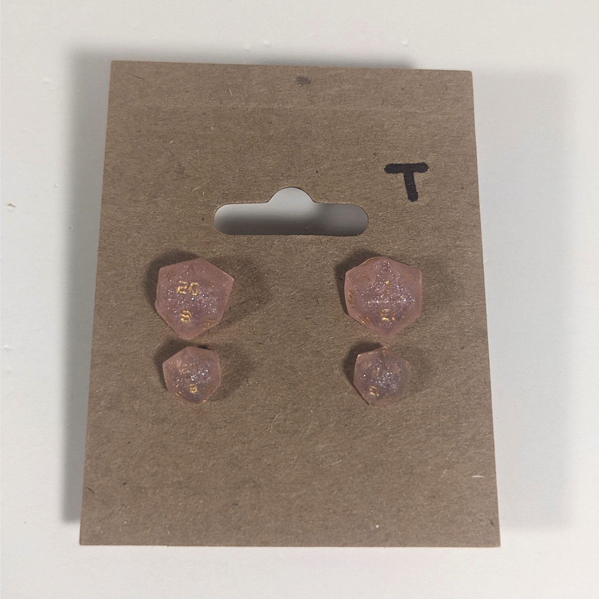 Two pairs of handmade d20 earrings with titanium hardware that are pastel pink with gold numbers on a kraft brown earring card with a T in the top right corner. The earrings on the left of each pair centers the 20 face and the earrings on the right of each pair centers the 1 face.