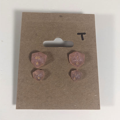 Two pairs of handmade d20 earrings with titanium hardware that are pastel pink with gold numbers on a kraft brown earring card with a T in the top right corner. The earrings on the left of each pair centers the 20 face and the earrings on the right of each pair centers the 1 face.