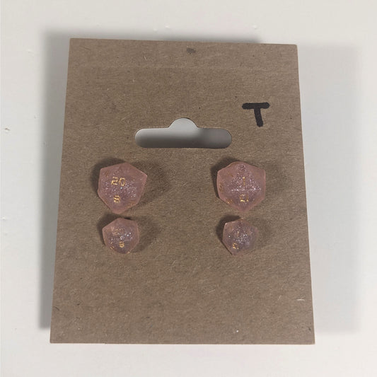 Two pairs of handmade d20 earrings with titanium hardware that are pastel pink with gold numbers on a kraft brown earring card with a T in the top right corner. The earrings on the left of each pair centers the 20 face and the earrings on the right of each pair centers the 1 face.