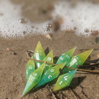 A vibrant set of green and yellow handmade dice resting on the sand of a sunny beach. The sharp diamond shape of these resin dice is clear against the coastal backdrop, with 2 d20s standing out among the other artisan dice in the collection.