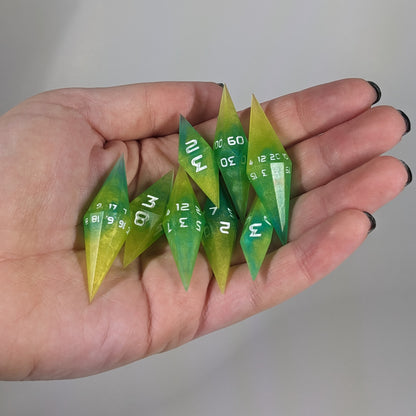Close-up of yellow and teal swirls with white numbers artisan dice resting in a palm on a seamless white backdrop. The handmade dice feature a distinct sharp diamond shape, with 2 d20s visible among the high-quality resin dice.