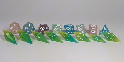 A size comparison of yellow and teal swirls with white numbers, long pointed diamond handmade dice with standard shapes placed behind for scale. The photo emphasizes the length of the diamond dice being much greater than a standard set as well as the diamond set being much more slender. This artisan dice set features 2 d20s and rests on a seamless white background to highlight the elongated profile of the resin dice.