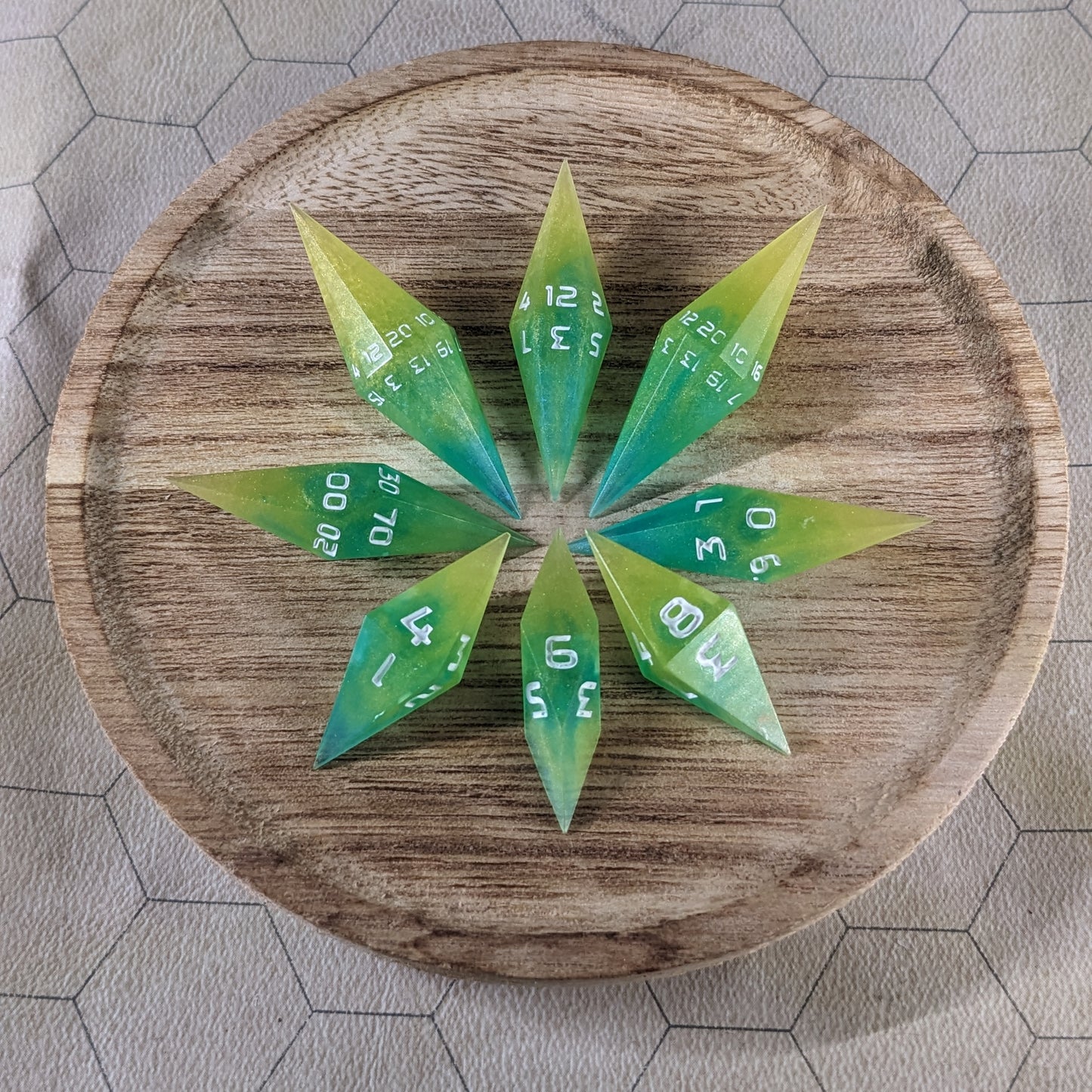 A set of yellow and teal swirls with white numbers handmade dice arranged in a wooden circle organizer. The resin dice feature a unique pointed diamond shape with the tips facing outward, highlighting the sharp edges of the two d20 and other artisan dice in the collection. 