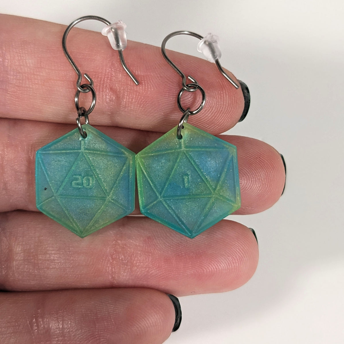 A pair of teal and yellow dangly, flat d20 earrings with silver color titanium and niobium hardware with bell shaped rubber backs held on on a hand on a white background. The earring on the left has a 20 on it and the earring on the right has a 1 on it, both in a sci-fi style font.