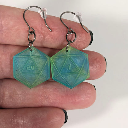 A pair of teal and yellow dangly, flat d20 earrings with silver color titanium and niobium hardware with bell shaped rubber backs held on on a hand on a white background. The earring on the left has a 20 on it and the earring on the right has a 1 on it, both in a sci-fi style font.