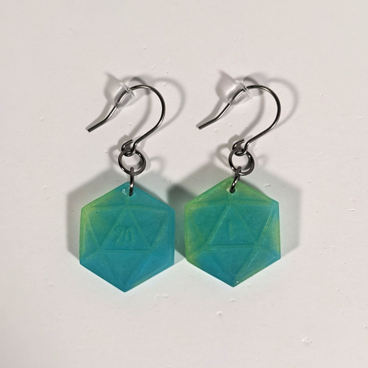 A pair of teal and yellow dangly, flat d20 earrings with silver color titanium and niobium hardware and bell shaped rubber backs on a white background. The earring on the left has a 20 on it and the earring on the right has a 1 on it, both in a fantasy style font.
