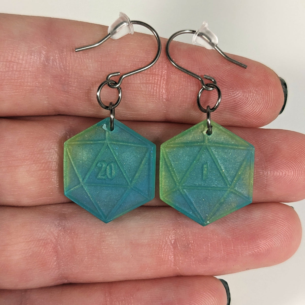 A pair of teal and yellow dangly, flat d20 earrings with silver color titanium and niobium hardware with bell shaped rubber backs held on on a hand on a white background. The earring on the left has a 20 on it and the earring on the right has a 1 on it, both in a fantasy style font.