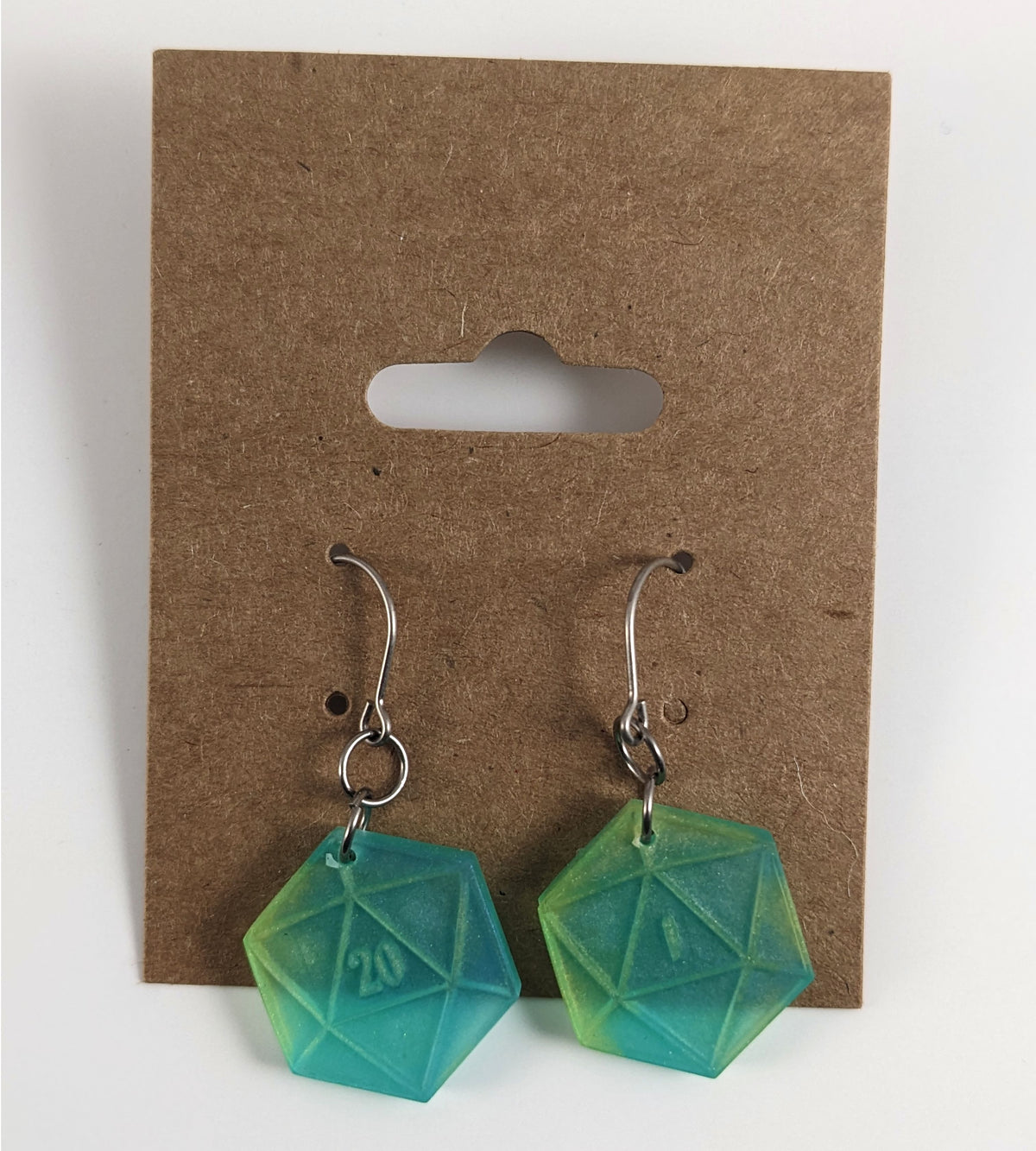 A pair of teal and yellow dangly, flat d20 earrings with silver color titanium and niobium hardware on a kraft earring card on a white background. The earring on the left has a 20 on it and the earring on the right has a 1 on it, both in a fantasy style font.