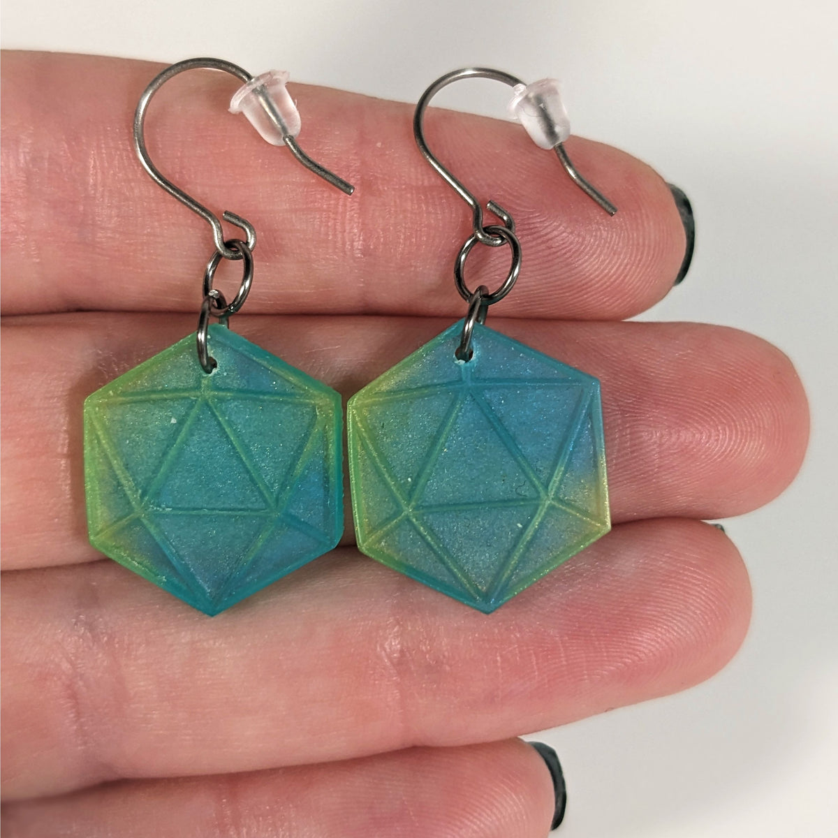 A pair of teal and yellow dangly, flat d20 earrings with silver color titanium and niobium hardware with bell shaped rubber backs held on on a hand on a white background.