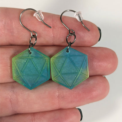 A pair of teal and yellow dangly, flat d20 earrings with silver color titanium and niobium hardware with bell shaped rubber backs held on on a hand on a white background.